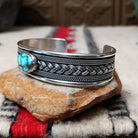 Silver cuff by H.M. featuring an oval blue turquoise stone with brown matrix, centered over an intricate braided silver band with twisted-wire detailing and oxidized accents; side view.