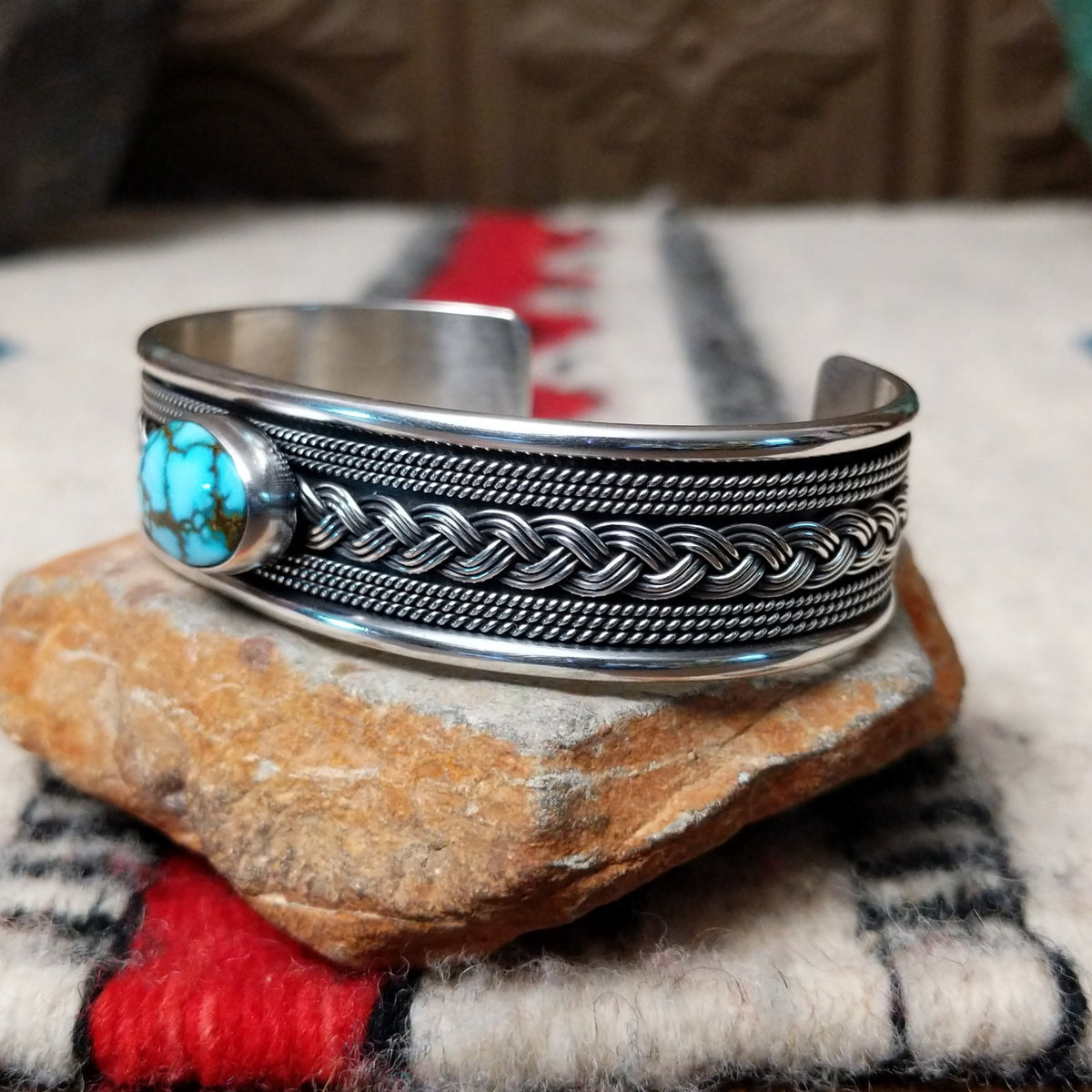Silver cuff by H.M. featuring an oval blue turquoise stone with brown matrix, centered over an intricate braided silver band with twisted-wire detailing and oxidized accents; side view.