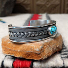 Silver cuff by H.M. featuring an oval blue turquoise stone with brown matrix, centered over an intricate braided silver band with twisted-wire detailing and oxidized accents; side view.