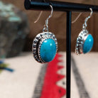 Turquoise earrings by Priscilla Smith featuring oval blue turquoise cabochons in decorative sterling silver settings with stamped borders and French wire hooks; side view.