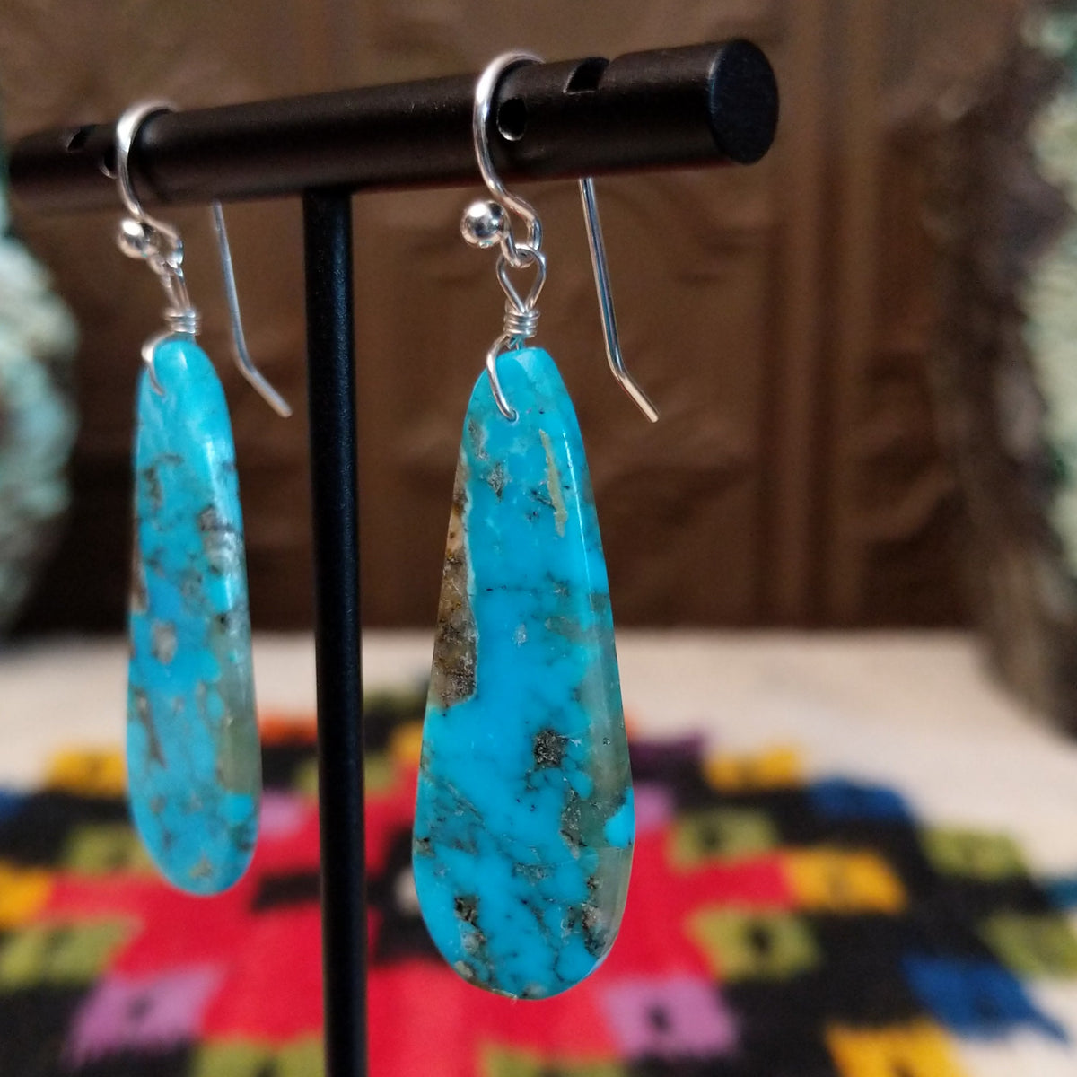 Turquoise Earrings by Ronald Chavez featuring elongated bright blue turquoise slabs with natural gray and brown matrix patterns, suspended from silver hooks; side view.