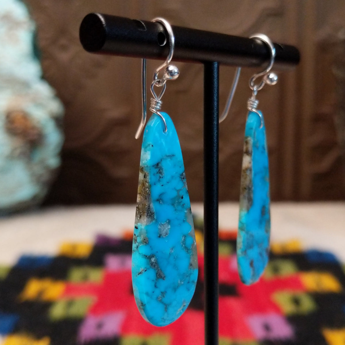 Turquoise Earrings by Ronald Chavez featuring elongated bright blue turquoise slabs with natural gray and brown matrix patterns, suspended from silver hooks; side view.