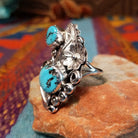 Turquoise Feather Ring by Harry B. Yazzie featuring two turquoise stones set in Sterling Silver with detailed feathers, a silver flower, scrollwork, and polished bead accents; side view.