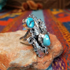 Turquoise Feather Ring by Harry B. Yazzie featuring two turquoise stones set in Sterling Silver with detailed feathers, a silver flower, scrollwork, and polished bead accents; side view.
