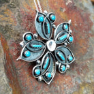 Handcrafted sterling silver turquoise flower pin and pendant by V. Booqua, featuring turquoise cabochons in a stylized floral design; side view.