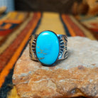 Turquoise Ring by Lowell Draper & Bernyse Chavez featuring a large oval turquoise stone with natural matrix, a polished silver bezel, and a wide silver band decorated with stamped sunburst patterns and geometric detailing.