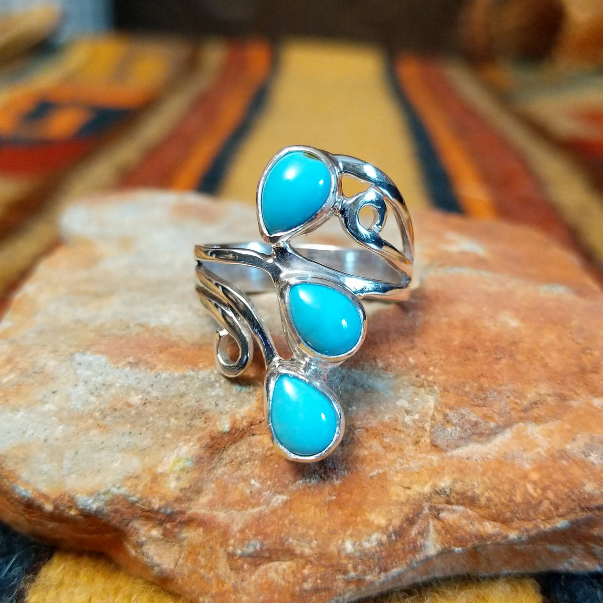 Front angled view of a silver ring by Raymond Coriz featuring three bright turquoise teardrop cabochons arranged vertically among flowing silver curves and spiral accents.