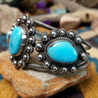 Handcrafted sterling silver cuff by Linda Johnson featuring three vibrant turquoise cabochons with twisted rope bezels, ornate silver beadwork, and a classic double-wire cuff design; side view.