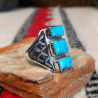 Turquoise Trio Ring by Pauline Nelson featuring three rectangular blue turquoise stones arranged vertically in serrated silver bezels, with detailed hand-stamped designs along the wide silver band; side view.