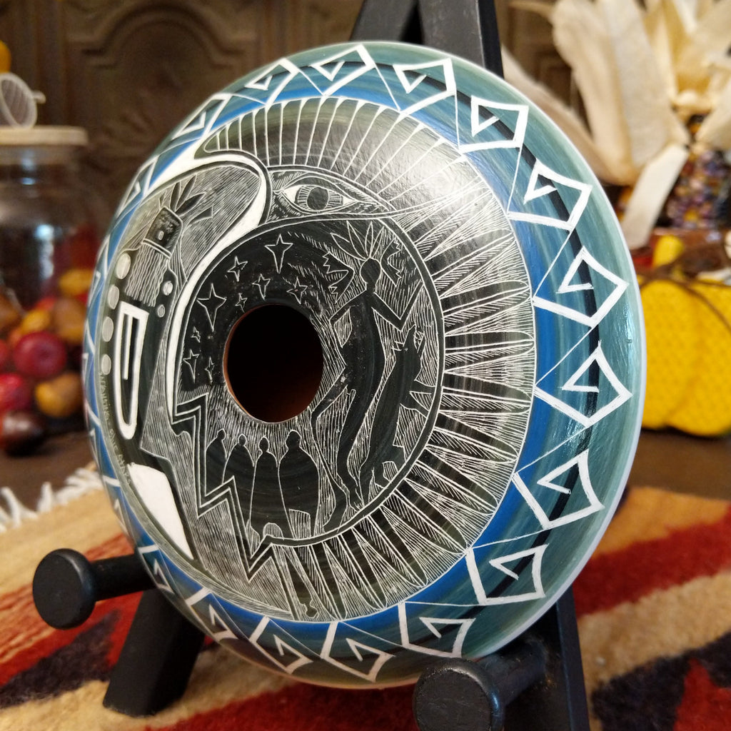 Clay with blue accents and carved with a spiritual figure, people, animals, and stars along the surface.