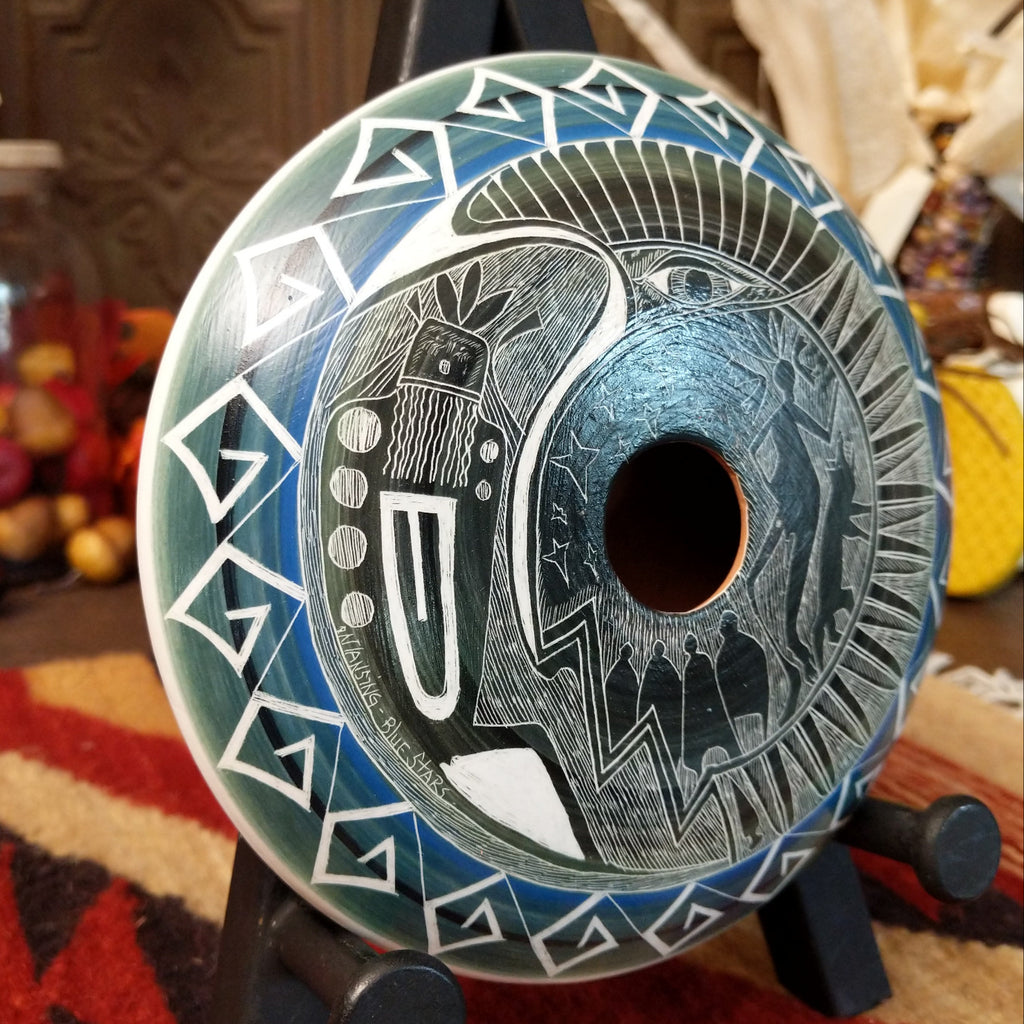Clay with blue accents and carved with a spiritual figure, people, animals, and stars along the surface.