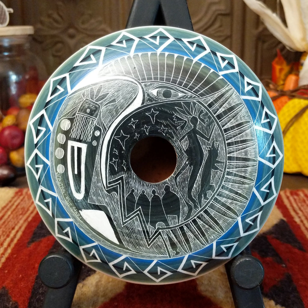 Clay with blue accents and carved with a spiritual figure, people, animals, and stars along the surface.