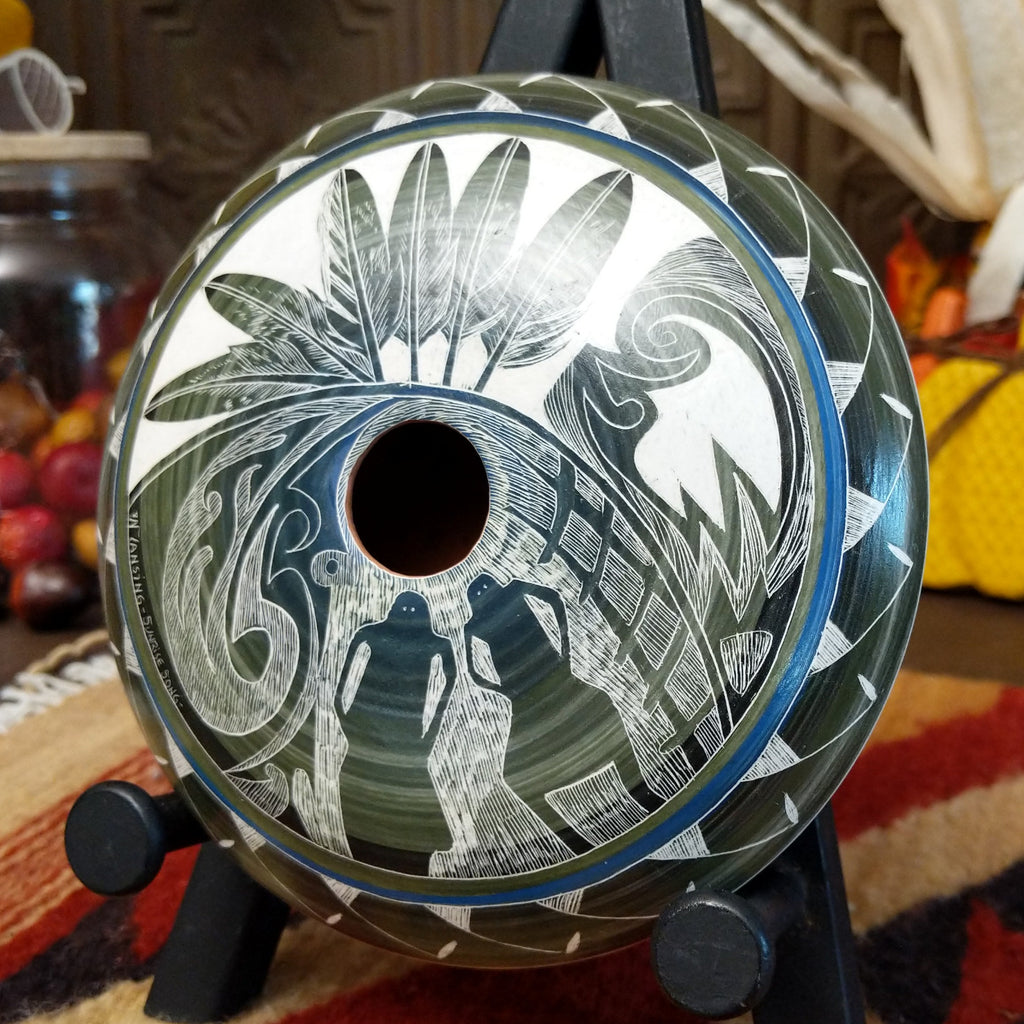 White clay with blue and green gradient and carved designs of people, feathers, and detailed patterns on the surface.