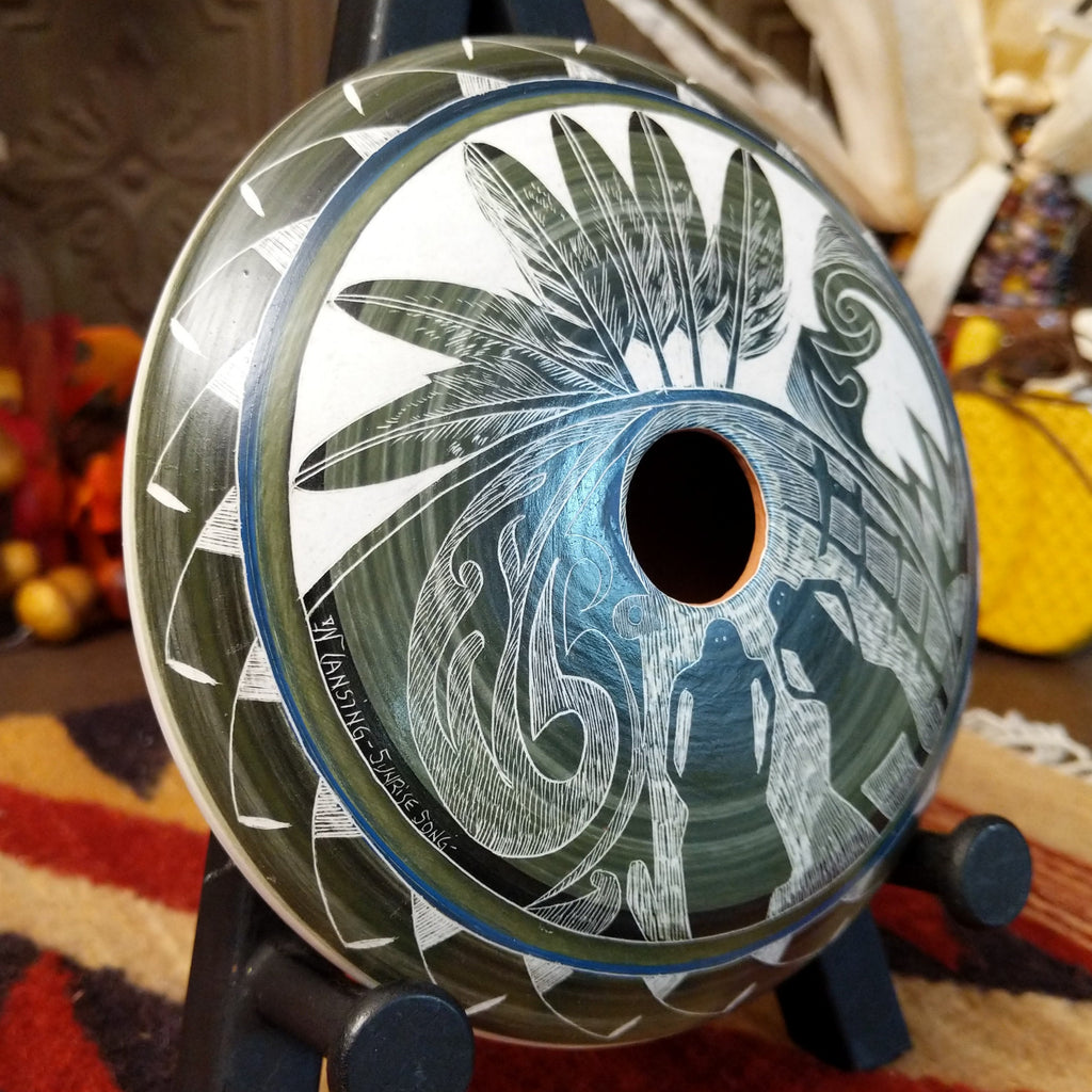 White clay with blue and green gradient and carved designs of people, feathers, and detailed patterns on the surface.