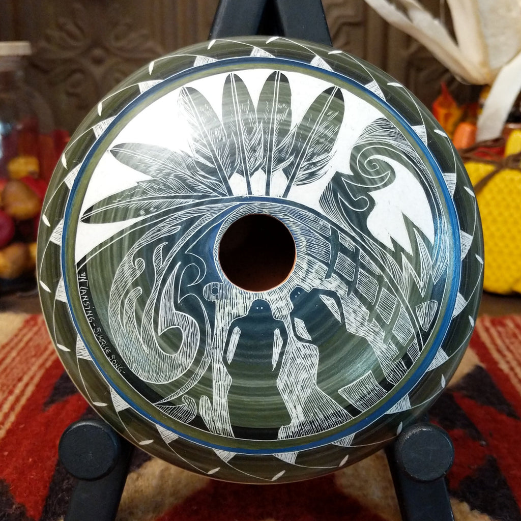 White clay with blue and green gradient and carved designs of people, feathers, and detailed patterns on the surface.