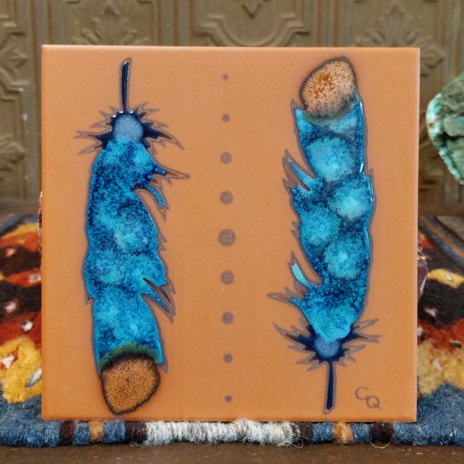 Ceramic tile featuring two blue watercolor style feathers with earthy tips on an orange background.

