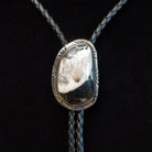 A silver-toned bolo tie necklace featuring a natural polished stone pendant with a white and dark marbled pattern, suspended on a braided dark cord against a black background.