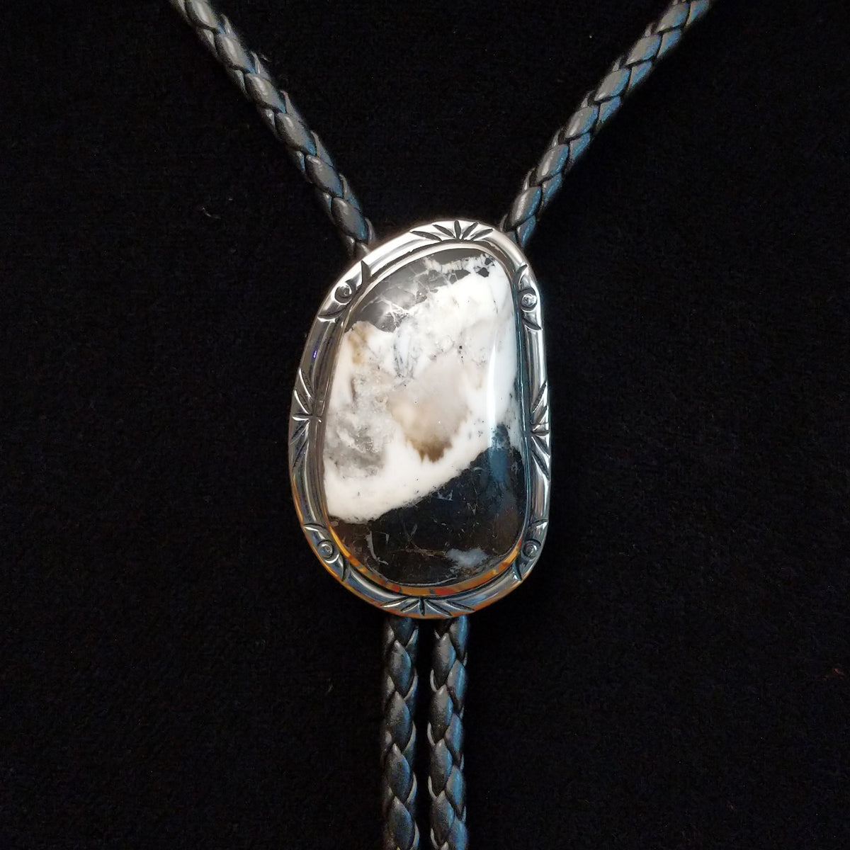 A silver-toned bolo tie necklace featuring a natural polished stone pendant with a white and dark marbled pattern, suspended on a braided dark cord against a black background.