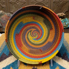 Handwoven wire art bowl by Bridge for Africa featuring a spiral pattern in recycled wire with olive green, mustard yellow, dusty blue, rust, terracotta, brown, and charcoal tones, perfect for home décor or wall display.