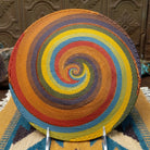 Handwoven wire art bowl by Bridge for Africa featuring a spiral pattern in recycled wire with golden yellow, olive green, dusty blue, rust, tan, brown, and charcoal tones, ideal for home décor or wall display; underside view.