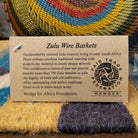 Handwoven wire art bowl by Bridge for Africa featuring a spiral pattern in recycled wire with olive green, mustard yellow, dusty blue, rust, terracotta, brown, and charcoal tones, perfect for home décor or wall display; artisan information.