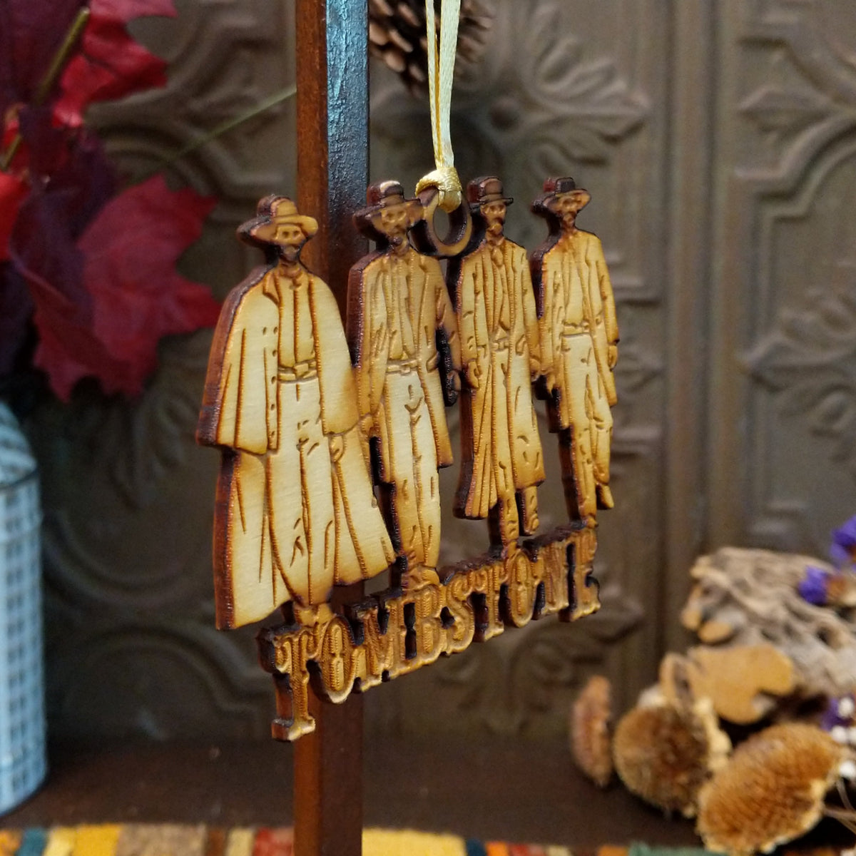 A wooden hanging ornament shaped like four cowboy figures with the word "TOMBSTONE" below them, suspended by a string in a styled indoor setting with autumn decor.
