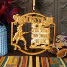 A gold-colored wooden Christmas ornament featuring the Tombstone 'The Town Too Tough to Die' design, hanging against a dark wooden background with rustic decorative props.
