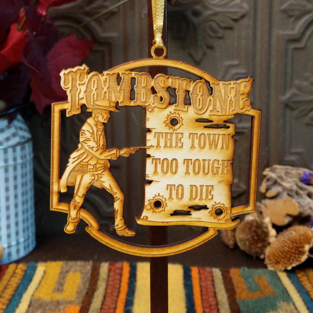 A gold-colored wooden Christmas ornament featuring the Tombstone 'The Town Too Tough to Die' design, hanging against a dark wooden background with rustic decorative props.