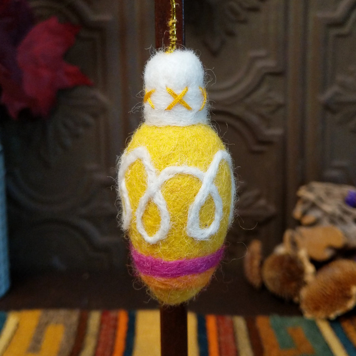 This delightful Yellow Bulb Wool Ornament by Ganz features a bright yellow wool body with white looped designs, a fluffy white top with yellow stitching, pink and orange bands near the bottom, and a gold hanging cord.