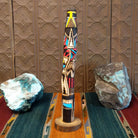 This distinctive Yellowface Shalako Kachina by John Taylor features a tall, slender wooden carving with a yellow face, intricate black geometric patterns, colorful red and turquoise blue accents, and a round wooden display base.
