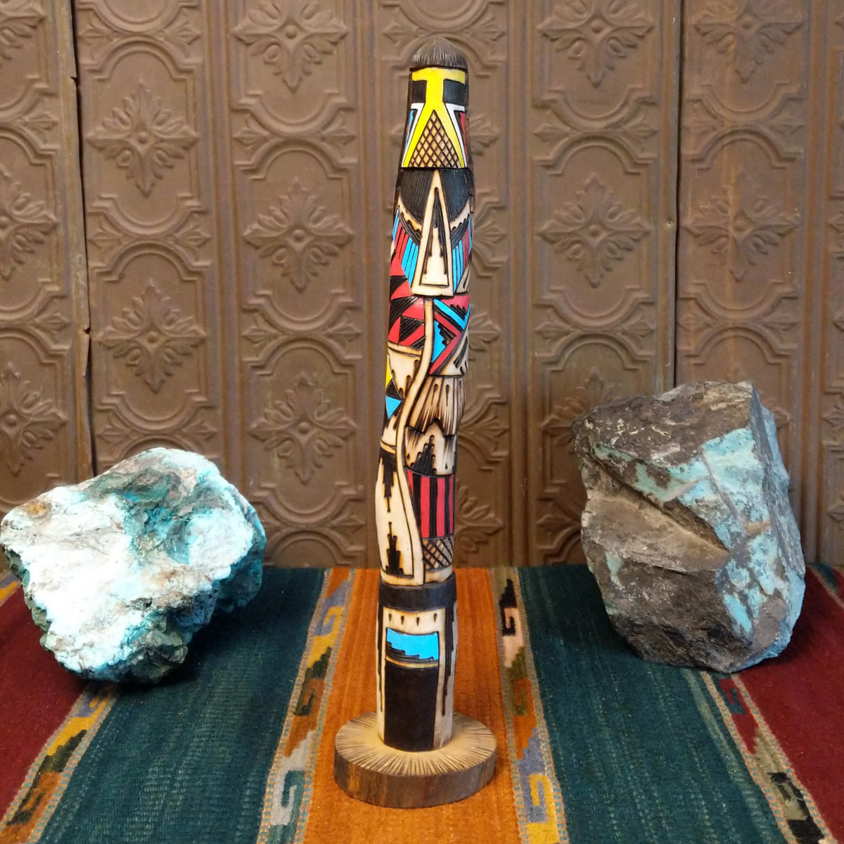 This distinctive Yellowface Shalako Kachina by John Taylor features a tall, slender wooden carving with a yellow face, intricate black geometric patterns, colorful red and turquoise blue accents, and a round wooden display base.