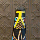 This distinctive Yellowface Shalako Kachina by John Taylor features a tall, slender wooden carving with a yellow face, intricate black geometric patterns, colorful red and turquoise blue accents, and a round wooden display base.