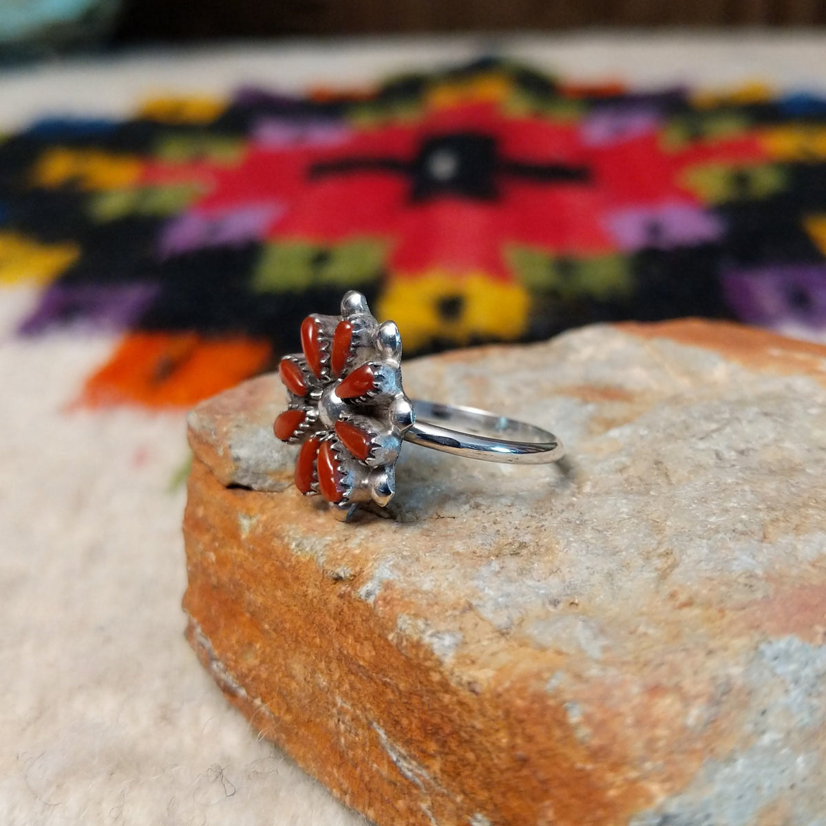 This vibrant Zuni Coral Flower Ring features teardrop-shaped red coral stones arranged as flower petals around a raised silver center, accented with rounded silver details; side view.