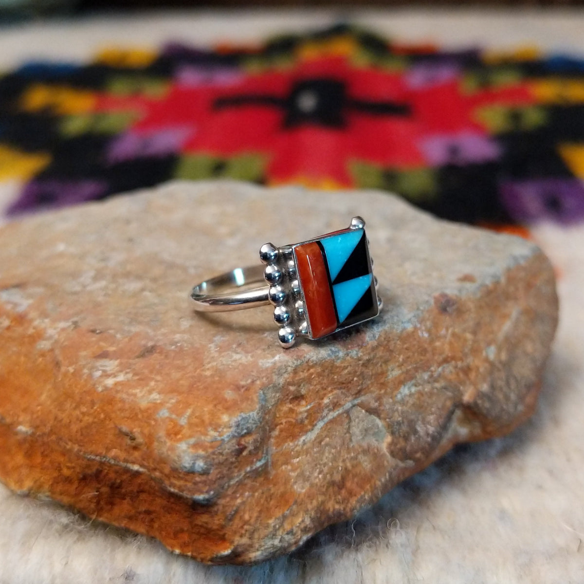 This colorful Zuni Inlay Ring features rectangular turquoise, black, and reddish-orange stone inlay in a geometric pattern, framed in silver with beaded accents along the sides; side view.