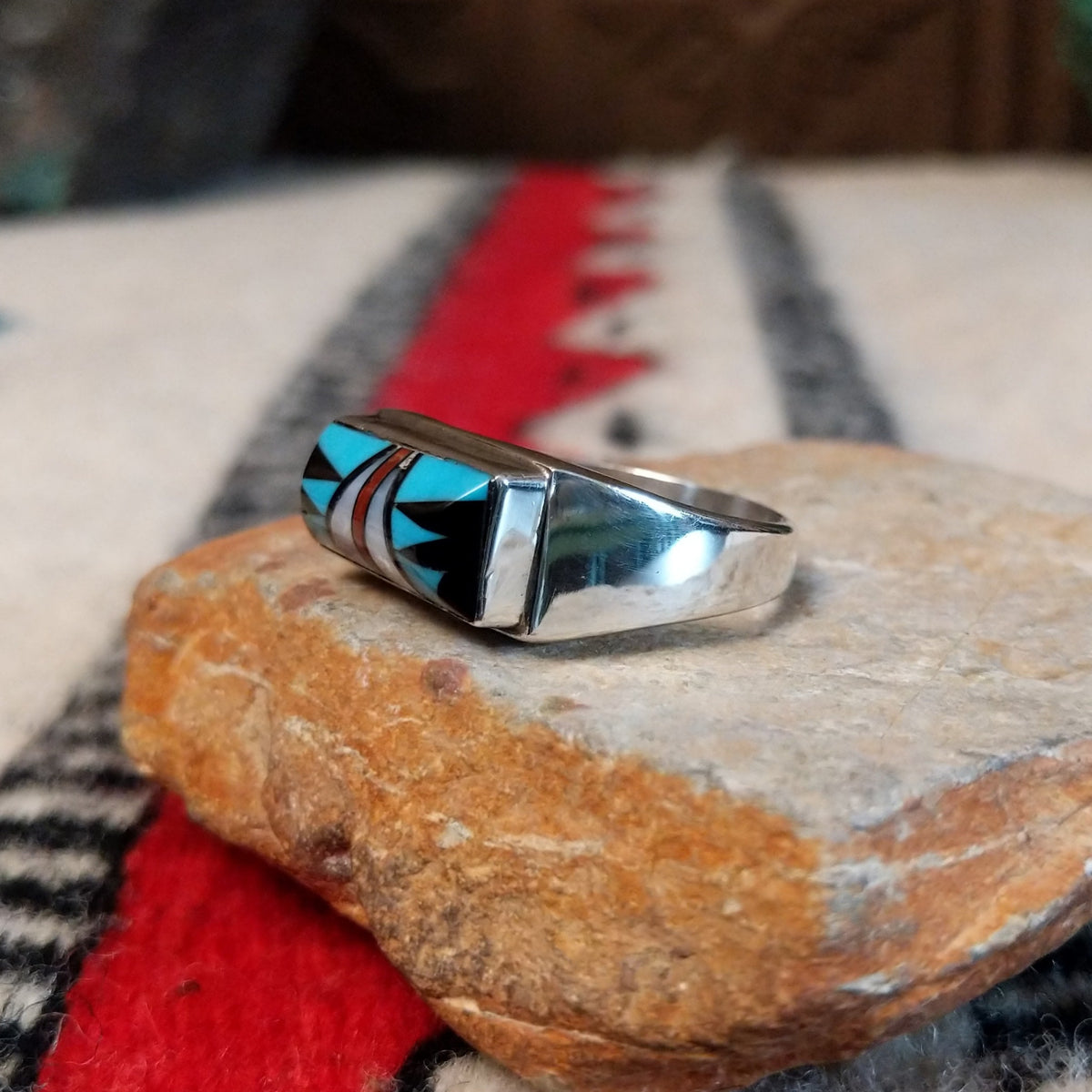 Zuni inlay ring by S.B. featuring a rectangular geometric design in turquoise, black onyx, coral, and mother of pearl inlay set in a polished silver band; side view.