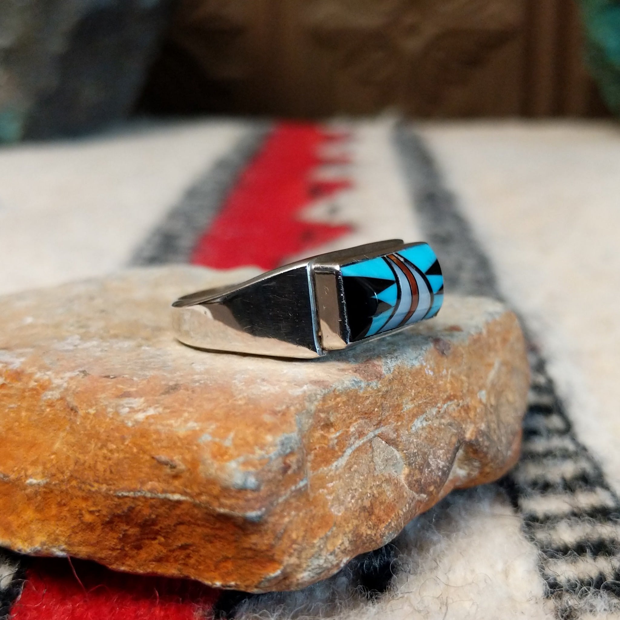 Zuni inlay ring by S.B. featuring a rectangular geometric design in turquoise, black onyx, coral, and mother of pearl inlay set in a polished silver band; side view.